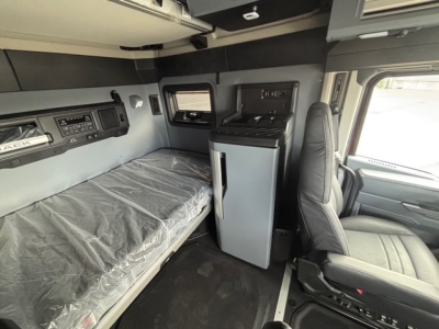Interior of a new 2026 Mack PR64T76H sleeper cab showing a single bed with plastic cover, cabinetry, control panel, and driver's seat.