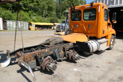 Orange Freightliner Cascadia Daycab truck with exposed drive line and missing cab interior showing chassis and drivetrain components.