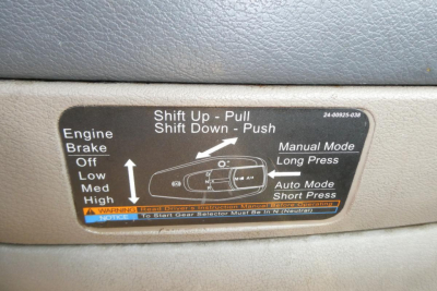 Truck interior control panel label showing shift instructions: 'Shift Up - Pull', 'Shift Down - Push', with diagrams and 'Manual Mode Long Press' and 'Auto Mode Short Press'. Also shows 'Engine Brake Off Low Med High' and safety warning tex