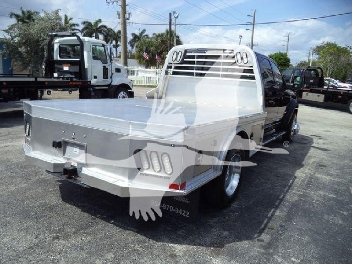 Flatbed truck rear view of a 2024 Ram 5500 SLT showing aluminum flatbed, custom rear frame, and twin vertical cab guards in a dealership lot.