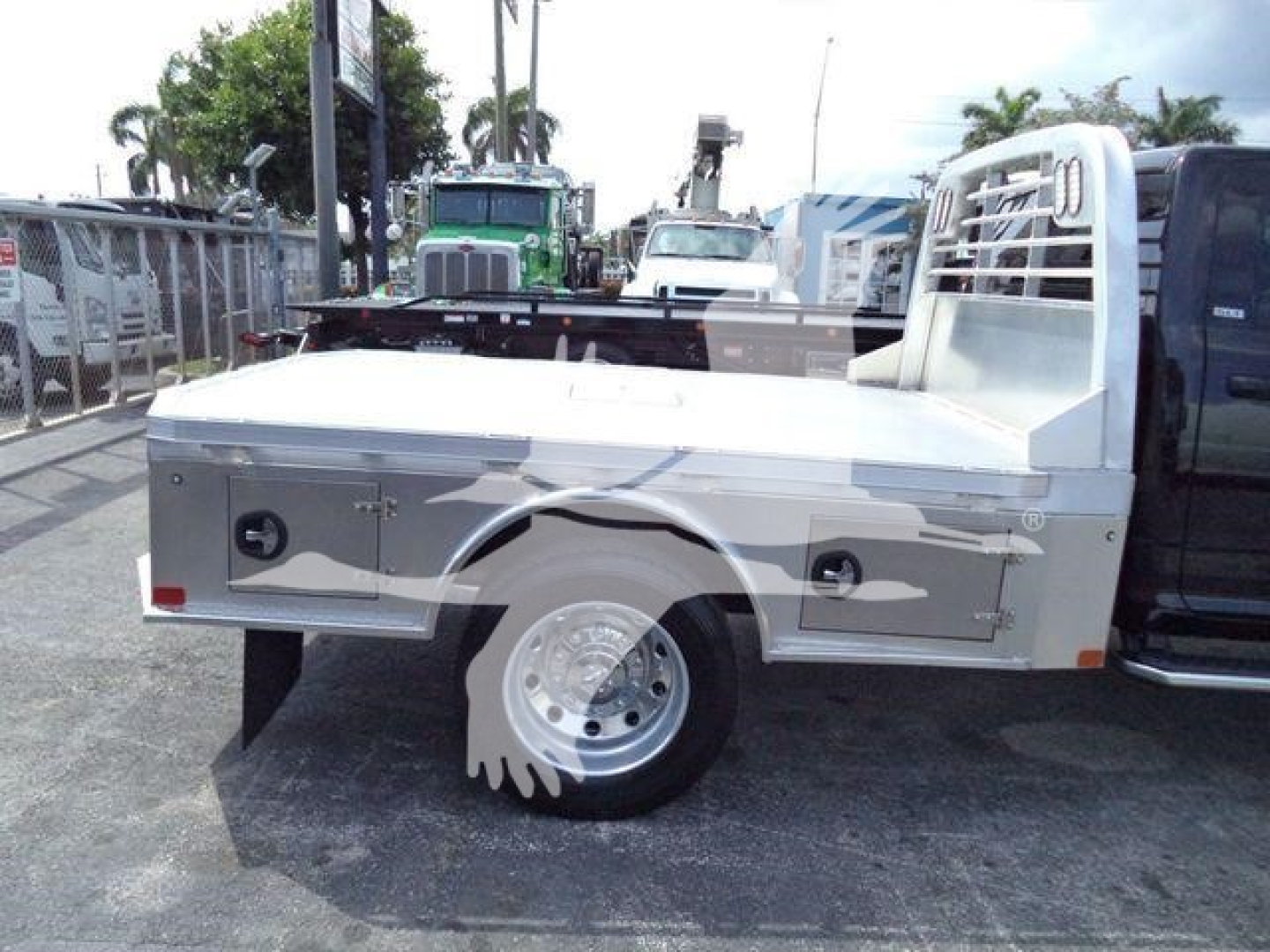 Flatbed truck side view showing storage compartments on silver tool boxes and a white wheel with a mud flap. Cab and bed with a protective cage behind cab.