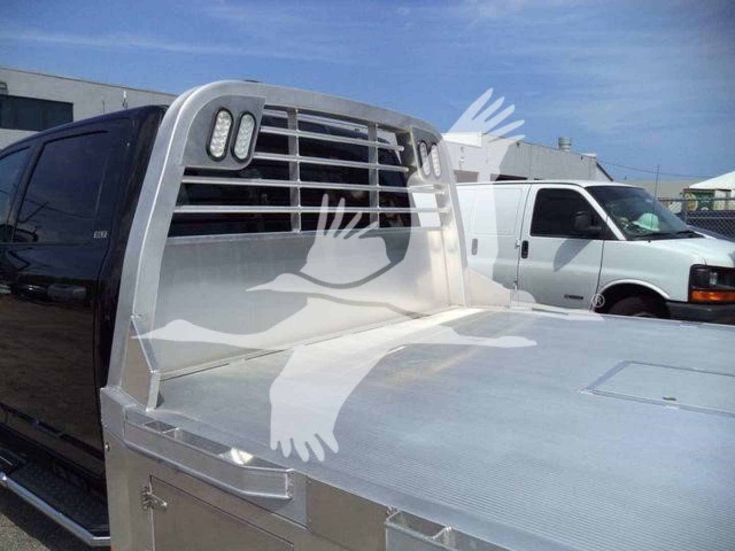 Ram 5500 flatbed truck with a silver headache rack and aluminum flatbed surface, grille and headlight area visible, other white cargo vans in background.
