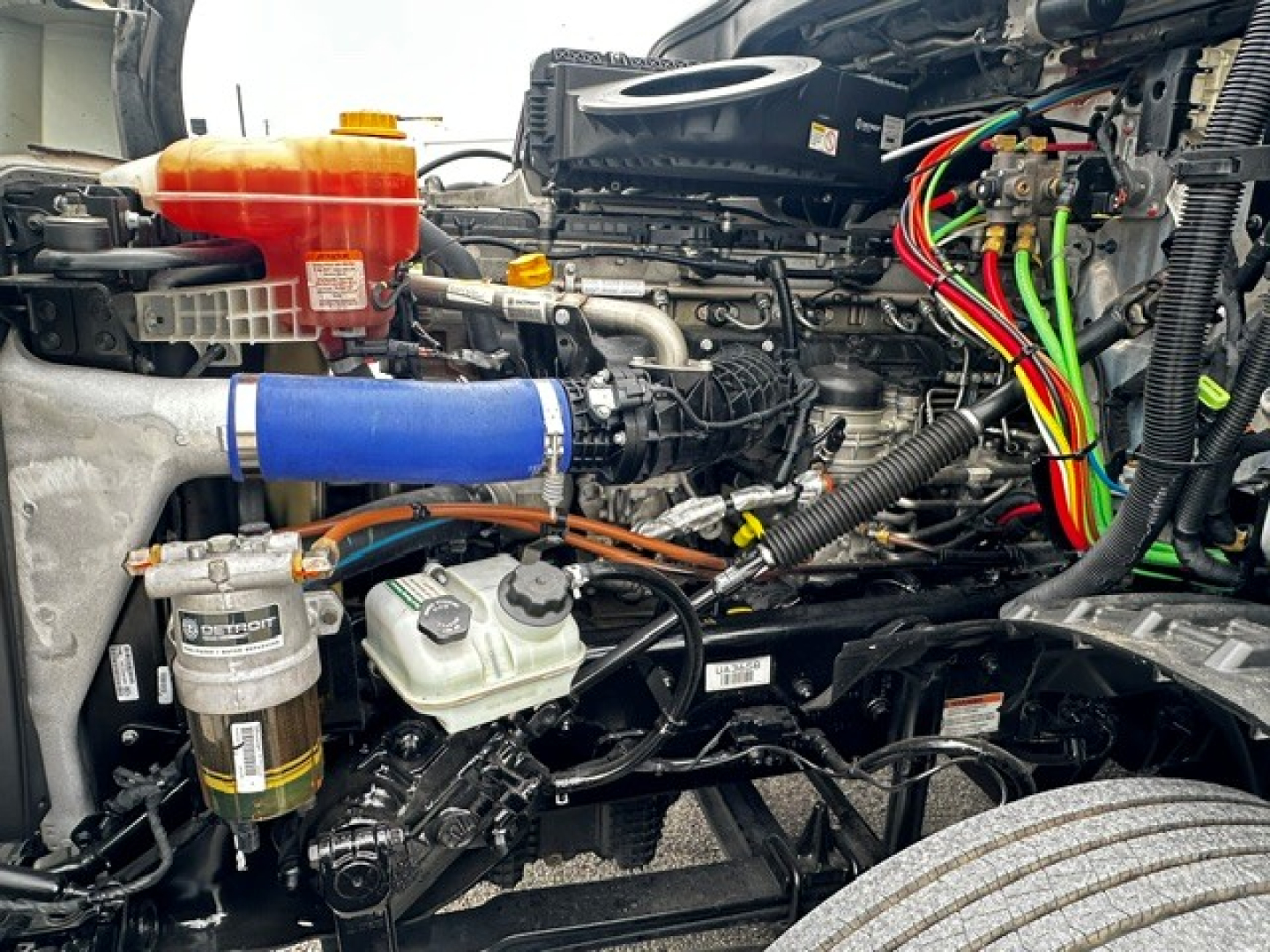 Truck engine bay showing various components including coolant reservoir (orange cap), blue intercooler pipe, hydraulic lines, and wiring harnesses.
