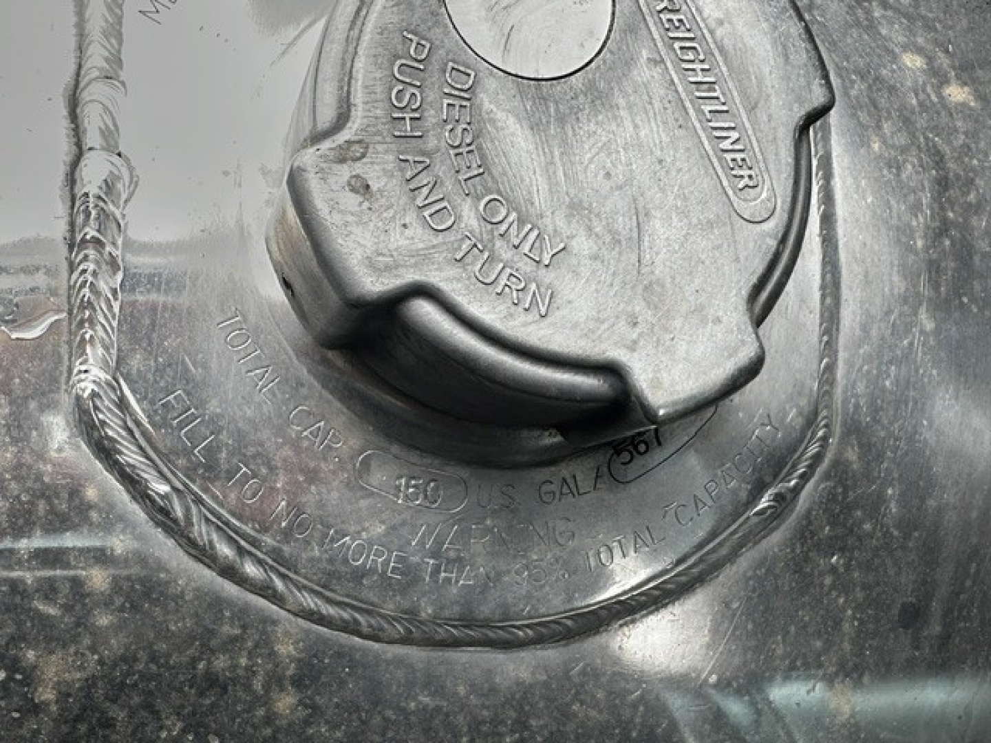 Fuel tank cap with warning text and cap outline visible in metallic surface; ribbed hose nearby.