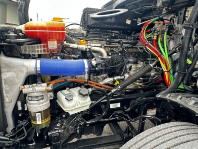 Truck engine bay showing various components including coolant reservoir (orange cap), blue intercooler pipe, hydraulic lines, and wiring harnesses.