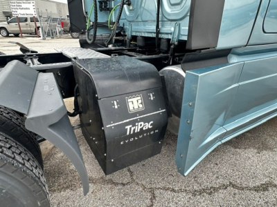 Truck side view showing black rectangular TriPac Evolution auxiliary power unit mounted behind cab on a blue Freightliner Cascadia 126.