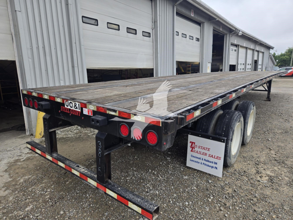 Flatbed trailer showing dual rear axles and wooden deck with reflective safety tape and rear lights.
