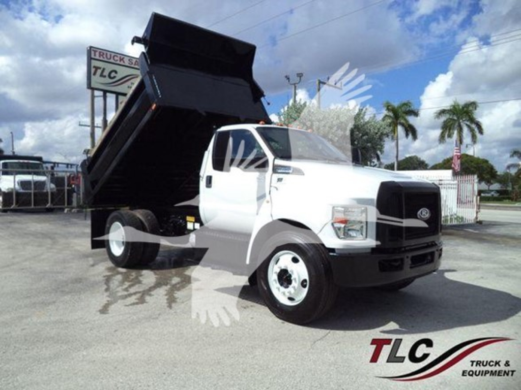 Dump truck in serviceable condition with raised dumping bed on a white Ford F-650, side view showing cab and front grille, dealership lot in background.