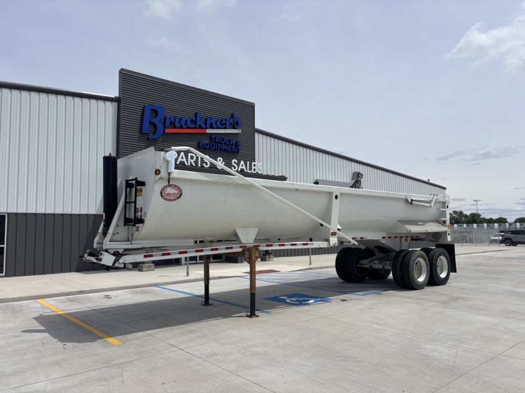 Dump trailer in a parking lot with an extended midsection and dual rear axles. Visible text includes a logo on the front of the trailer and the Bruckner sign on the building.