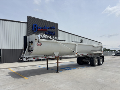 Dump trailer in a parking lot with an extended midsection and dual rear axles. Visible text includes a logo on the front of the trailer and the Bruckner sign on the building.