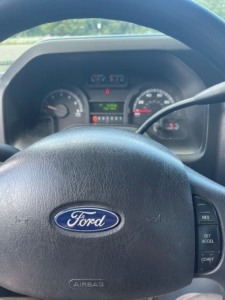 Ford steering wheel with instrument cluster in background displaying gauges and trip controls. Partial dashboard visible.