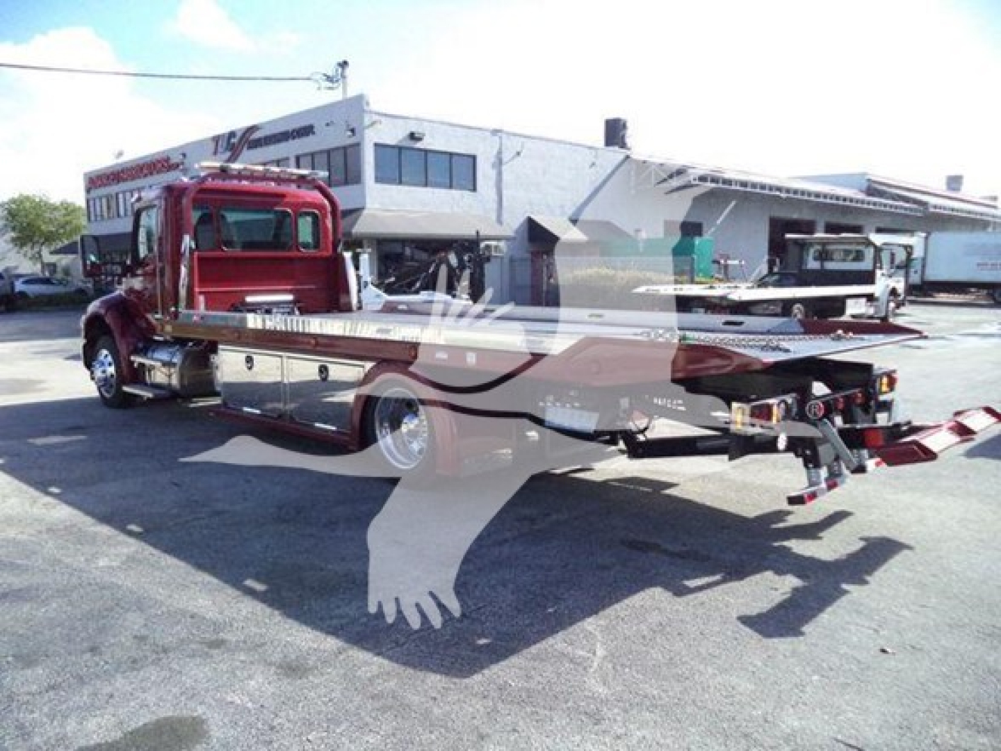 Kenworth rollback truck with a long flatbed and hydraulic winch, red cab, rear wheel lift, and vehicle dealership lot in background.