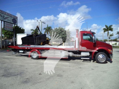 Kenworth T280 rollback tow truck side view showing red cab and flatbed with equipment on bed and black toolbox; dealership lot in background.