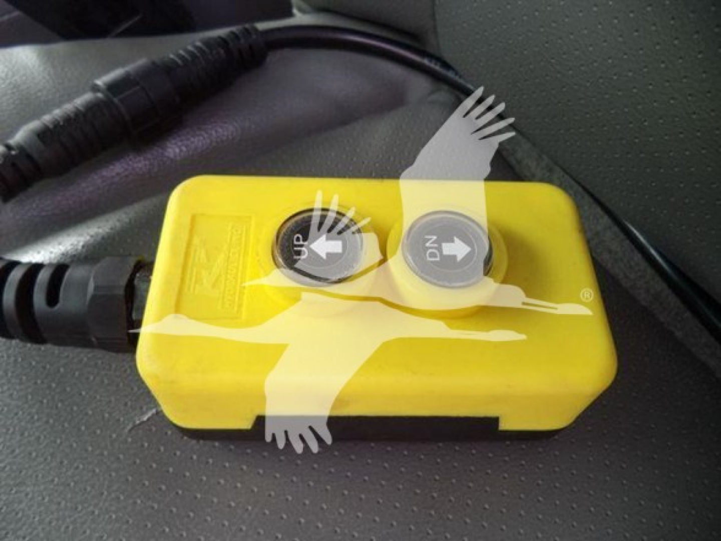 Yellow hand-held device with two gray circular dials labeled UP and DN, connected by a black cord, placed on a vehicle seat.