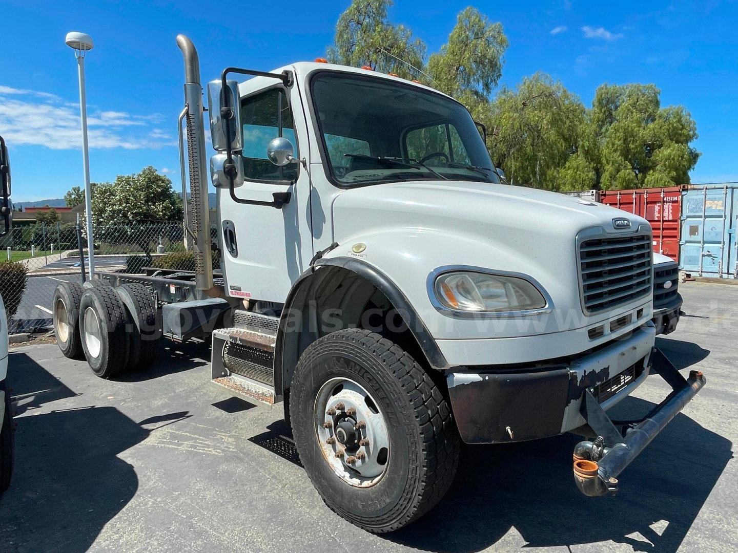 Truck cab and chassis showing Freightliner M2 106 front view with dual rear axles, white cab, black bumper, and chrome exhaust stack.