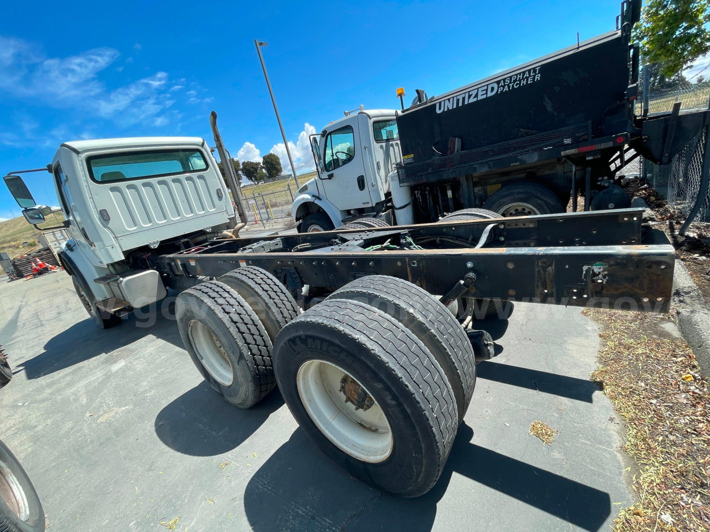Truck chassis with dual rear axles and cab in background. Visible text on truck includes 'UNITIZED DUMP' on black dump truck in background and the 2009 Freightliner M2 106 listing details in the context; no legible alphanumeric engine specs