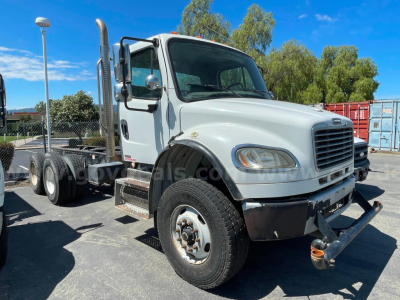 Truck cab and chassis showing Freightliner M2 106 front view with dual rear axles, white cab, black bumper, and chrome exhaust stack.