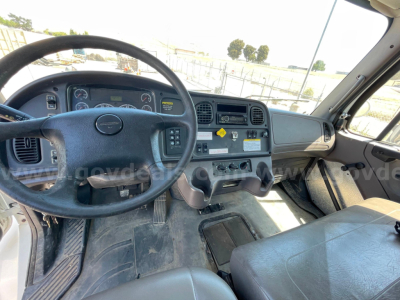 Cab interior of a Freightliner M2 106 showing steering wheel, dashboard, center console, and driver seat from driver’s side. Includes gauges, vents, and control knobs.