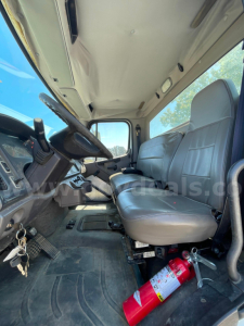 Cab and chassis interior showing driver and passenger seats, steering wheel, pedals, and a fire extinguisher on the floor near the seat. Visible headliner and door frame.
