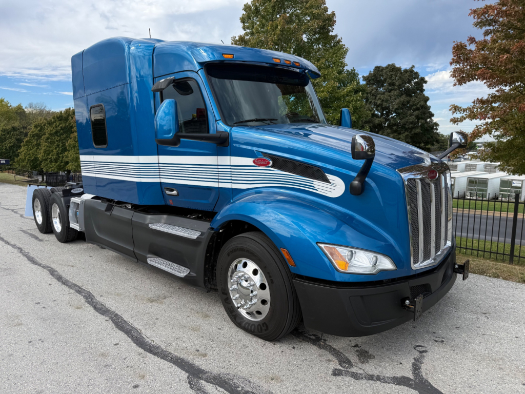 Peterbilt 579 truck in blue with sleeper cab, chrome grille, white stripes along the side, front view showing headlights and dual rear tandem wheels.
