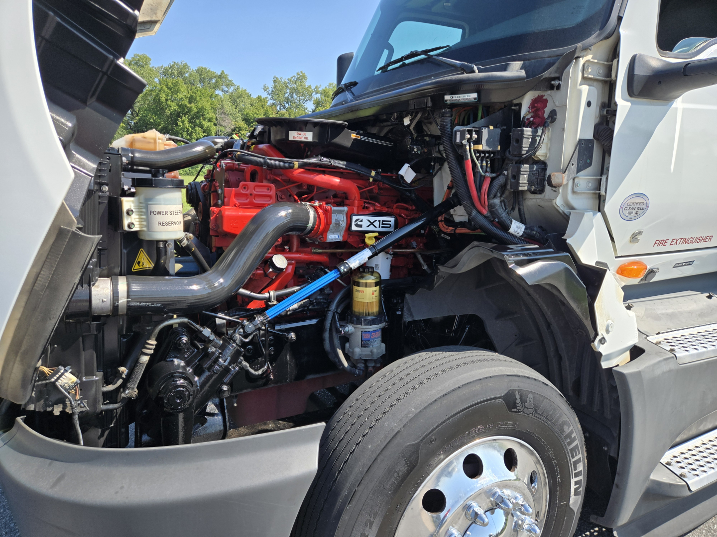 Truck engine bay of a 2021 International LT625 with red engine, black hoses, and nearby silver wheel; visible components include coolant reservoir, air intake, and various hoses.