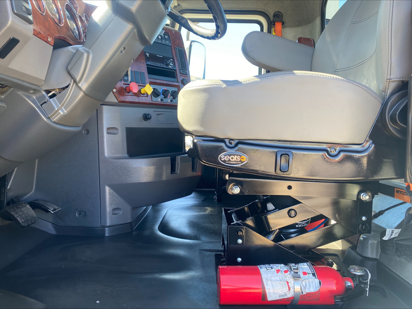 Cab interior of a 2018 Mack CXU612 truck showing driver's seat with Seats logo, steering wheel, dashboard controls, and a mounted fire extinguisher under the seat.