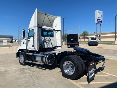 Truck cab and chassis of a 2018 Mack CXU612 with open hood and exposed engine area, dual rear axles, chrome wheels, and white cab. Visible signs in background.