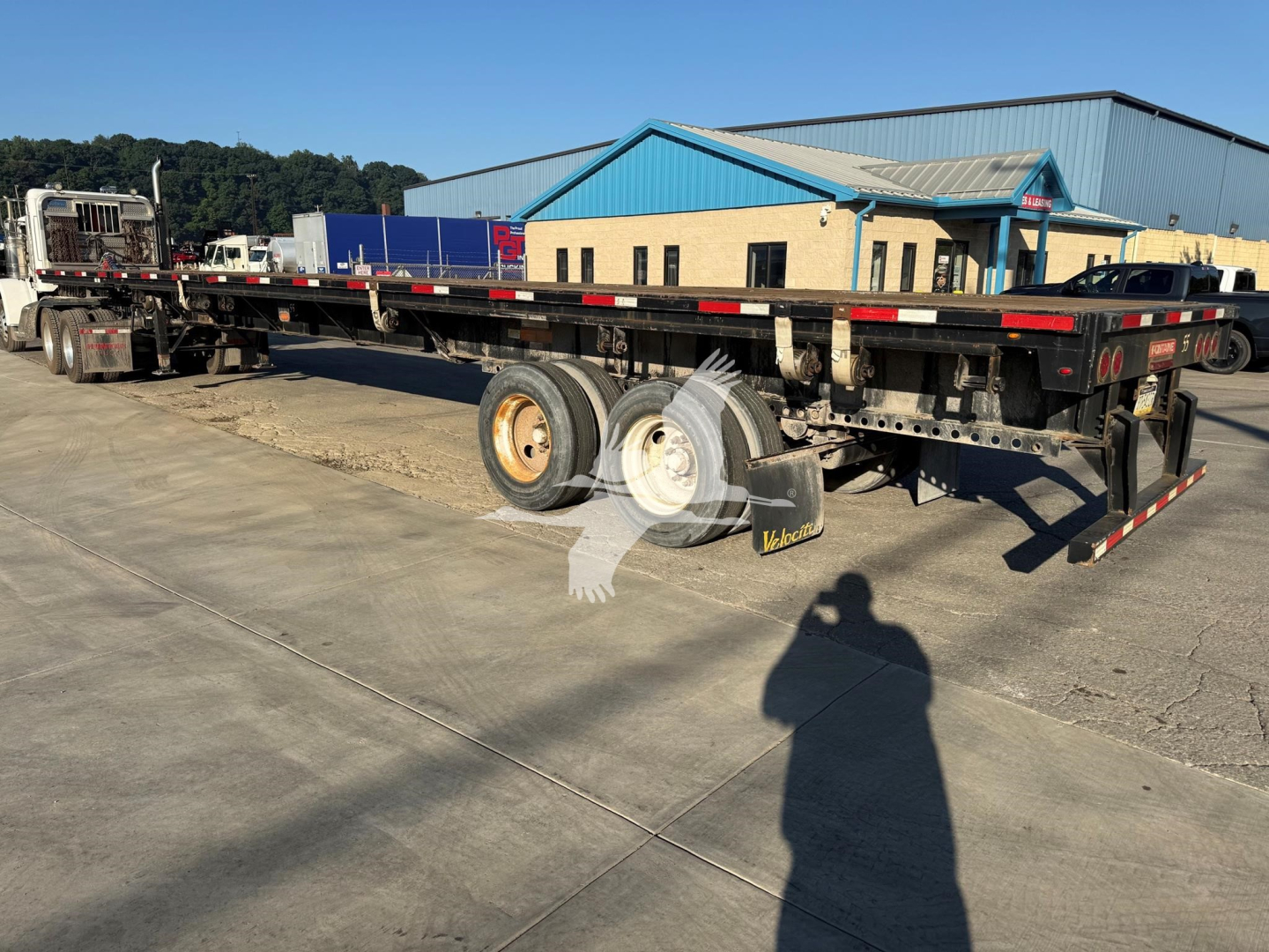 Flatbed trailer in service showing long main deck, tandem axle setup, and wheel chocks with Vox logo in foreground. Text and model details not legible.