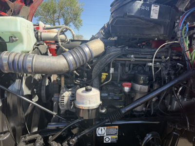 Kenworth T880 engine bay showing air intake hose, coolant reservoir, oil filter, and various engine components in a daytime setting.