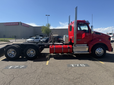 Kenworth T880 conventional daycab truck side view showing red cab, dual rear axles, fuel tanks, chrome exhaust stacks, and chassis in a parking lot.