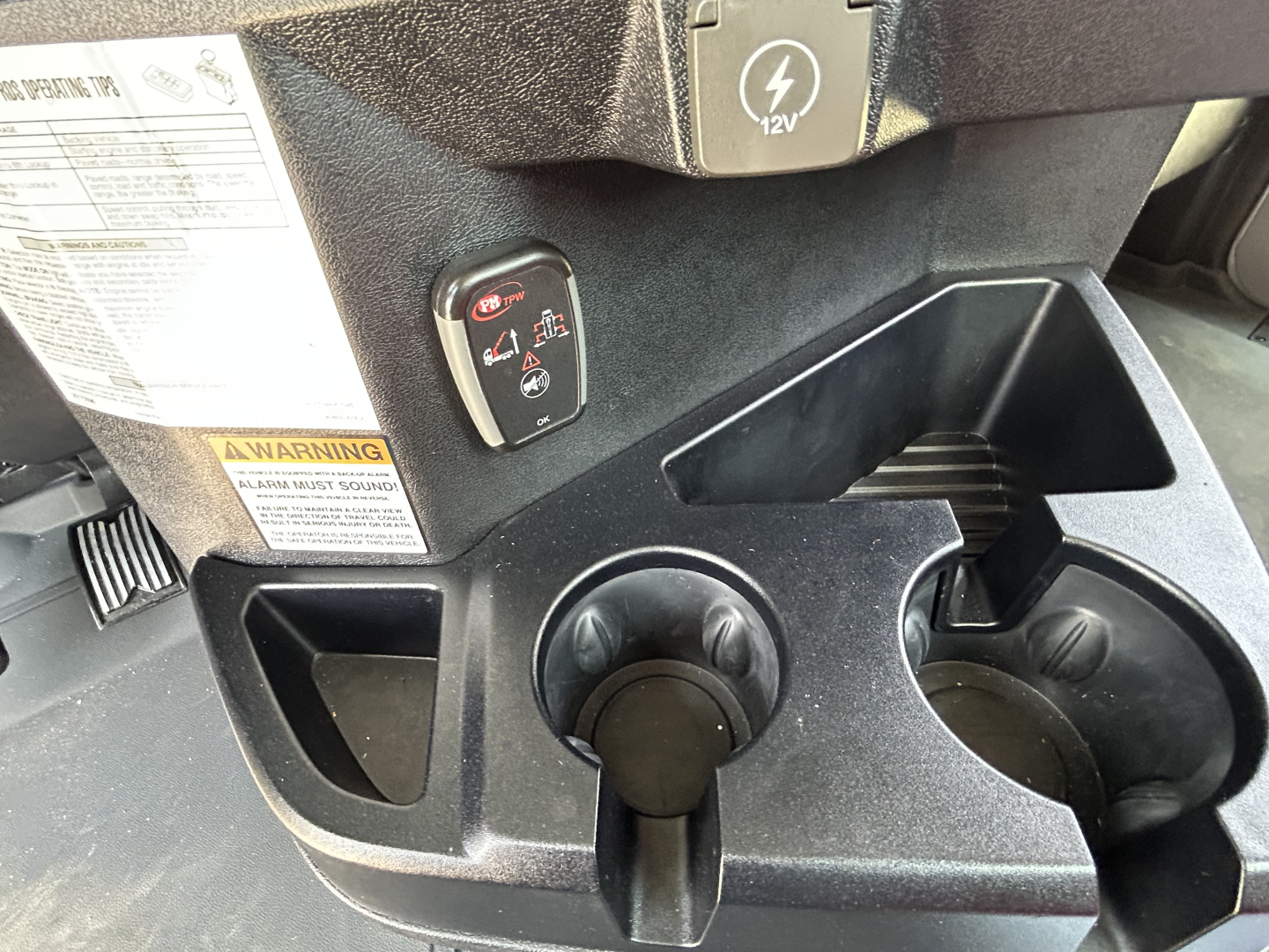 Dashboard area of a new Mack GR64F crane truck showing a 12V power outlet, cup holders, and small switches with labels.
