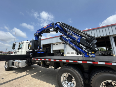 Flatbed tow truck with blue hydraulic crane mounted on chassis, Mack GR64F crane model, side view showing crane arms folded over bed, white cab.