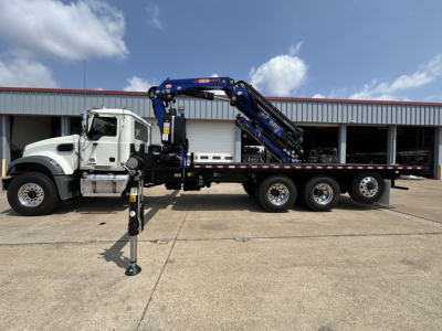 Truck with a mounted crane (GR64F) on a long flatbed, side view showing stabilization outriggers extended, Mack cab, blue crane arm, black chassis, six wheels on rear, industrial yard background.