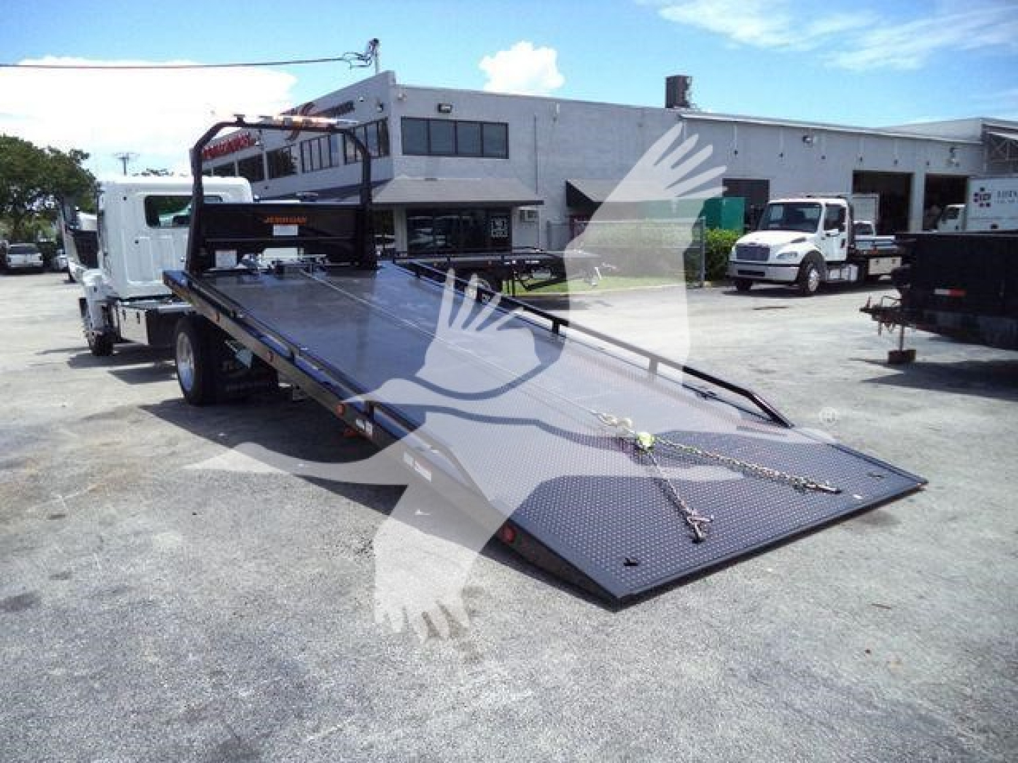 Flatbed tow truck with long ramp and winch, loading area visible, Hino L6 2027 new unit in lot with other trucks in background.