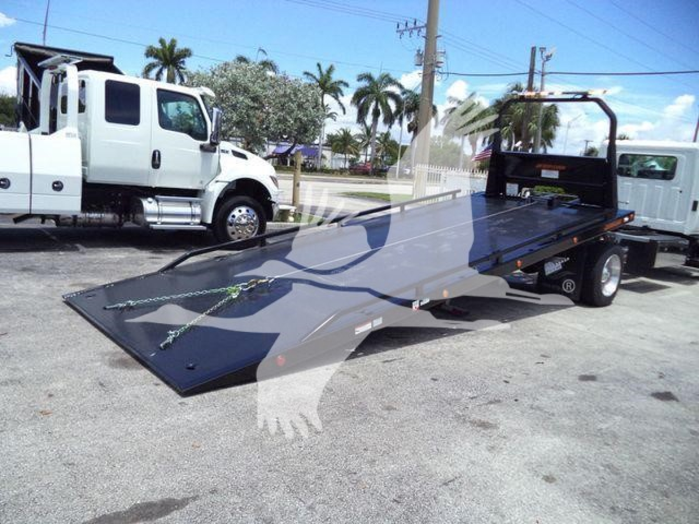 Flatbed tow truck with a low-profile steel ramp in a parking lot, white cab in background, chain tie-downs visible on the ramp, Hino branding not visible.