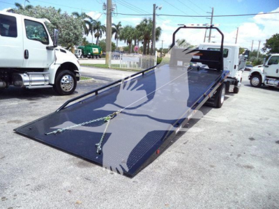 Flatbed rollback truck with a lowered black steel ramp bed, white cab, and a winch and straps visible on the ramp; background shows other trucks and palm trees.