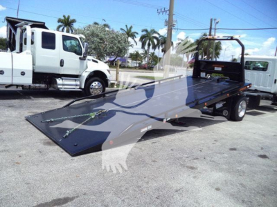 Flatbed tow truck with a low-profile steel ramp in a parking lot, white cab in background, chain tie-downs visible on the ramp, Hino branding not visible.