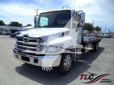 Hino L6 rollback truck in white, front three-quarter view showing chrome grille and flatbed with equipment in yard.