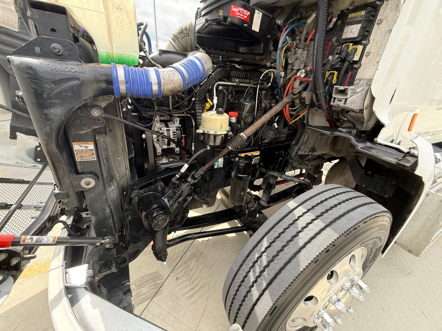 Peterbilt 579 engine bay with visible hoses, wiring, and front suspension. Cylinders and components visible near radiator and air intake; blue hose wrap on intake; multiple cables and connectors.