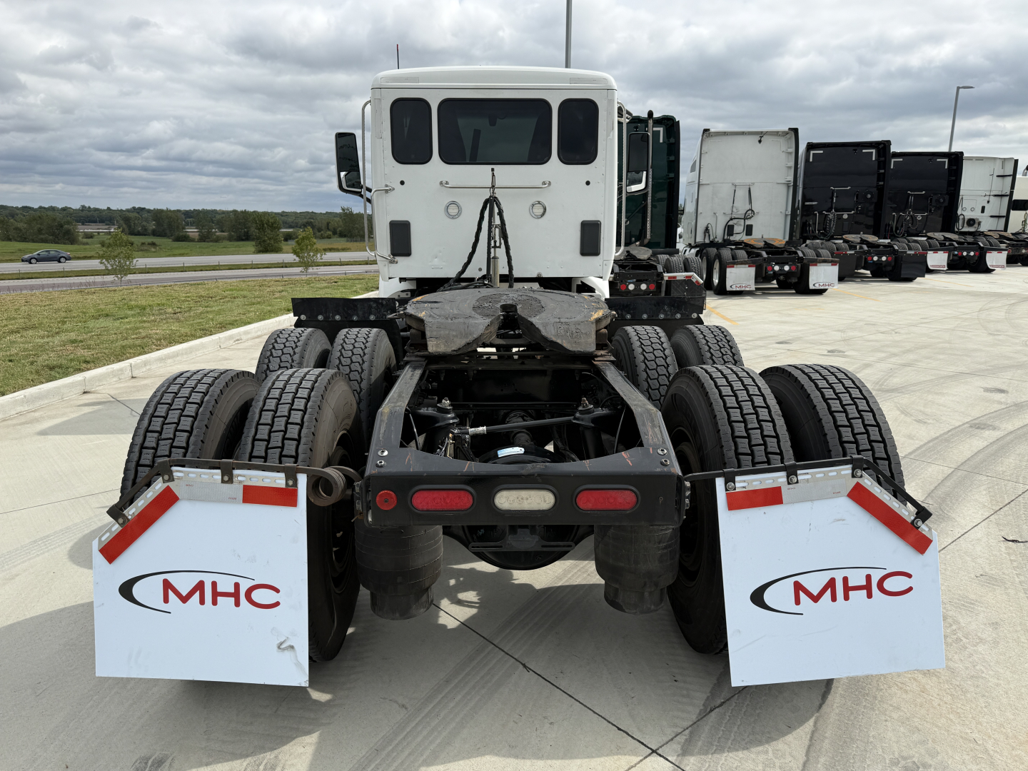Peterbilt 579 rear chassis and dual rear axles with mud flaps showing MHC logos, flatbed area, and fifth-wheel; white cab in distance.