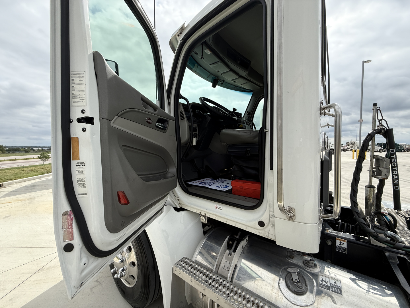 Peterbilt 579 truck cab interior with open door; dashboard and steering wheel visible; steps and chassis near doorway; some labels on door frame.