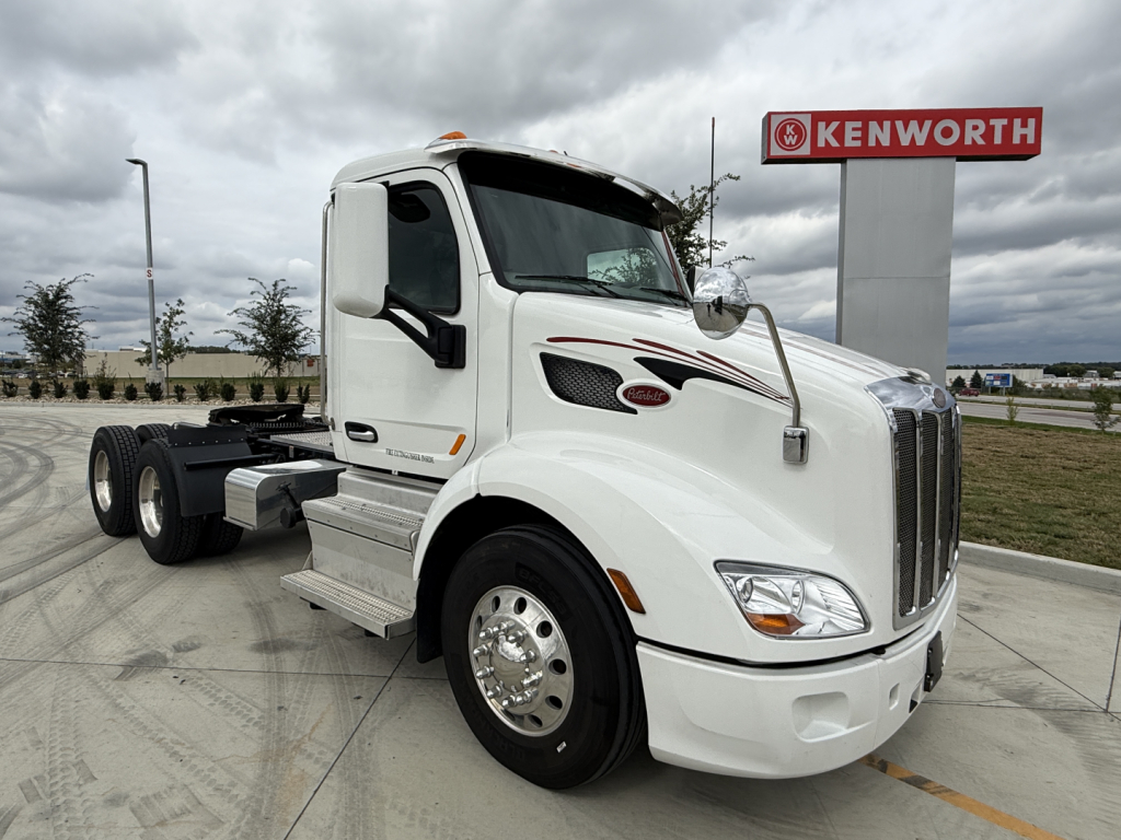 Peterbilt 579 tractor cab in white with Kenworth sign in background, front left view showing grille, headlights, and side mirrors.