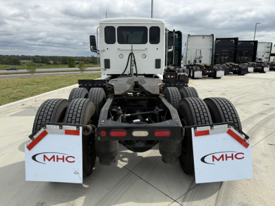 Peterbilt 579 rear chassis and dual rear axles with mud flaps showing MHC logos, flatbed area, and fifth-wheel; white cab in distance.