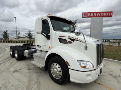 Peterbilt 579 tractor cab in white with Kenworth sign in background, front left view showing grille, headlights, and side mirrors.