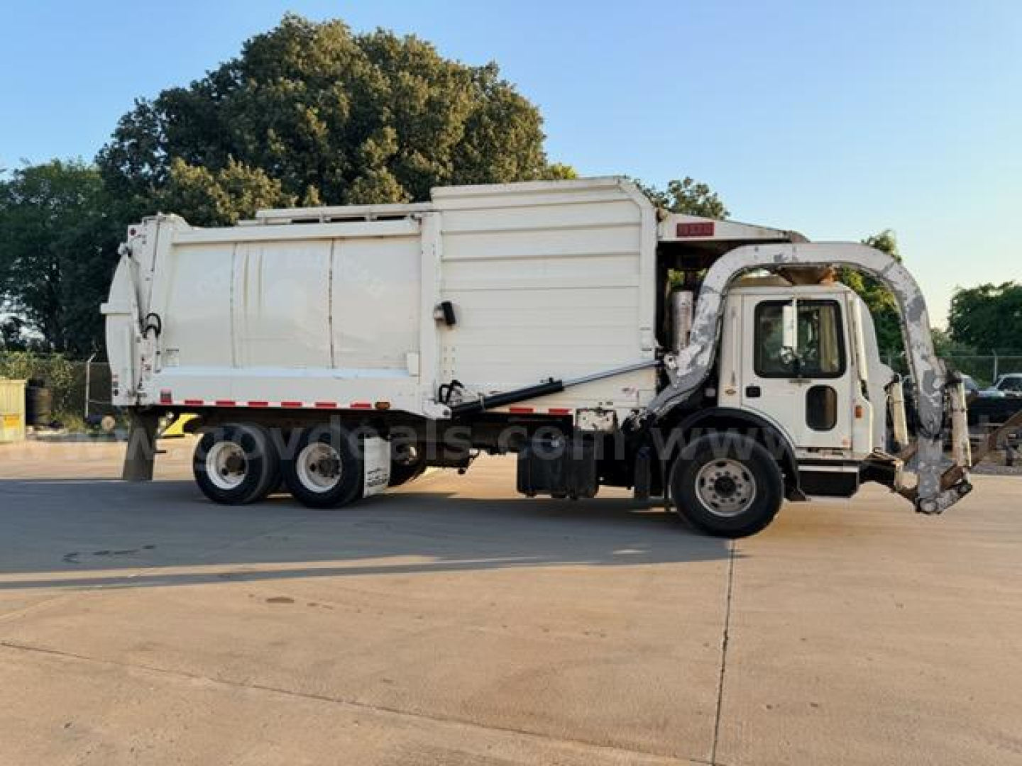 Truck side view of a white 2018 Mack MRU613 refuse truck with bin body and side loader, dual rear axles, and cab with open doorway.