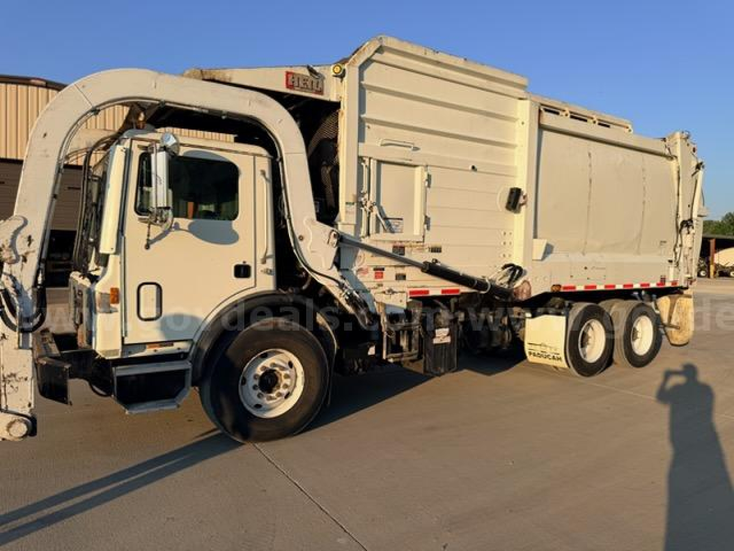 Mack MRU613 dump truck in beige with front tow device and large side compactor, cab visible, multiple wheels, VIN 1M2AV04C3JM019254 not visible on plate.