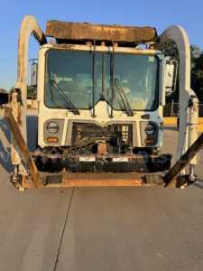 Front view of a 2018 Mack MRU613 garbage truck cab with metal arms on sides and large windshield; rust and wear visible on front frame.