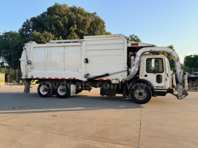 Truck side view of a white 2018 Mack MRU613 refuse truck with bin body and side loader, dual rear axles, and cab with open doorway.