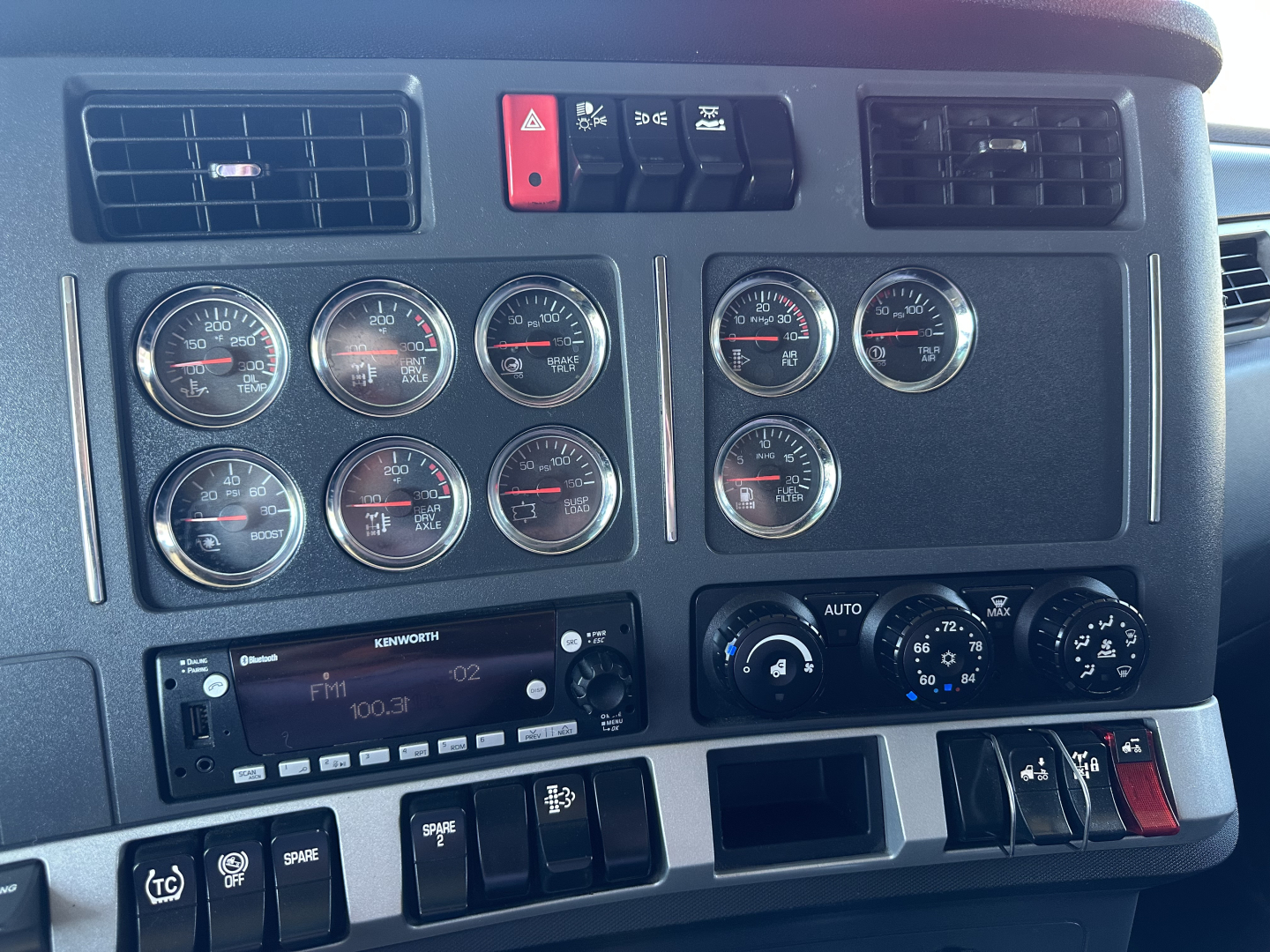 Dashboard of a Kenworth truck showing multiple round gauges, switches, and a Kenworth audio/CD player. Labels visible include Infiniti-style dials and climate controls.