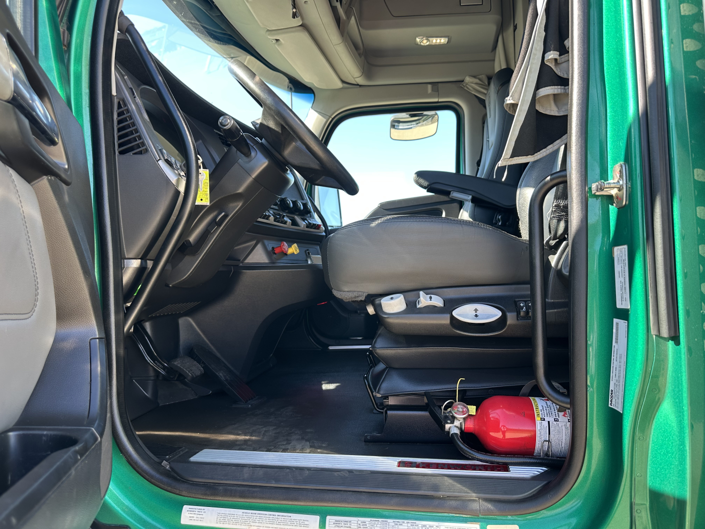 Kenworth T680 truck interior driver seat and dashboard view from open door showing steering wheel, ignition area, and fire extinguisher in cab.